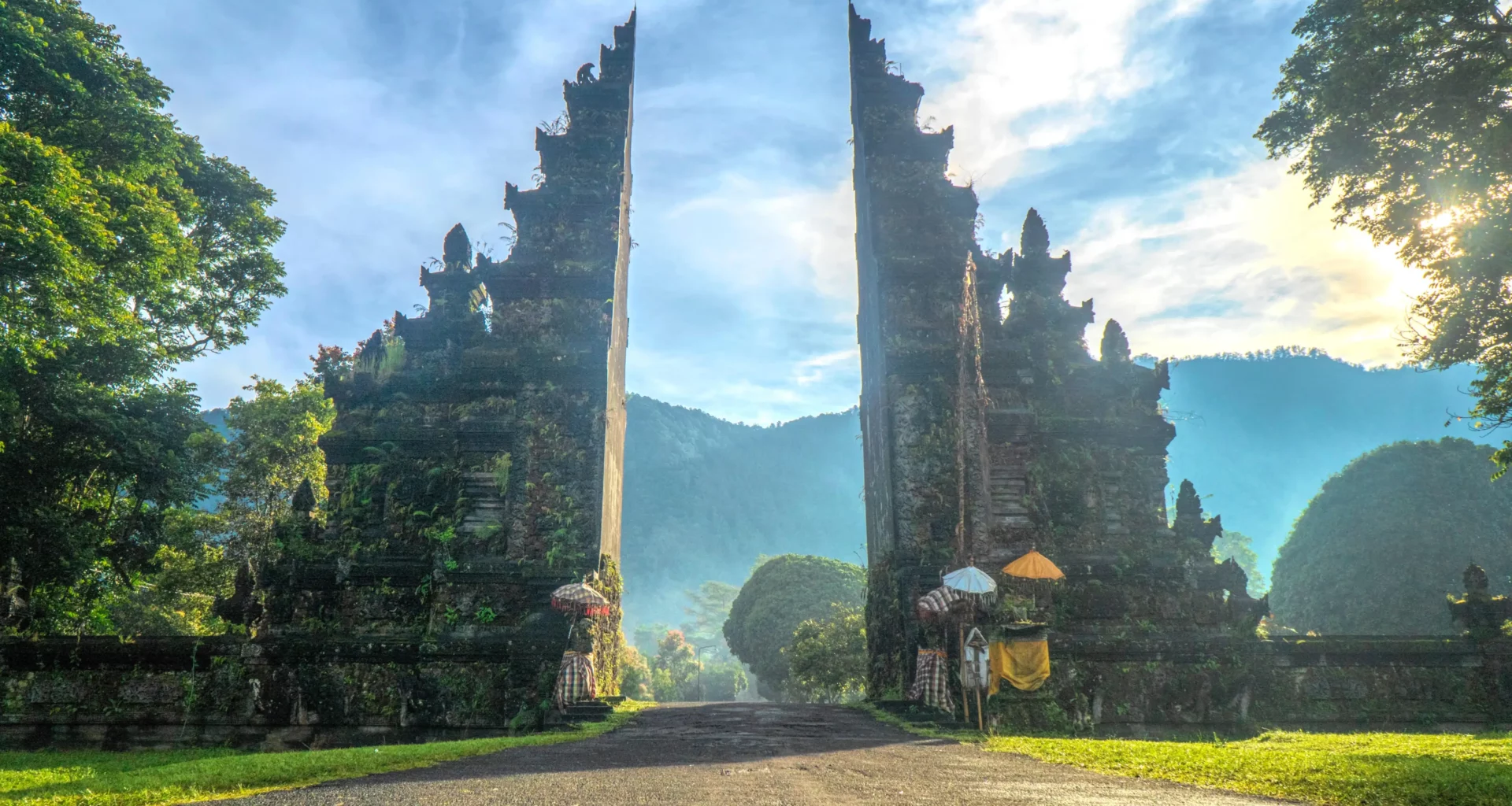 Entrance to a luxury resort in Bali, with traditional Balinese gate structures, lush green landscape, and a serene, tropical backdrop under a bright blue sky.