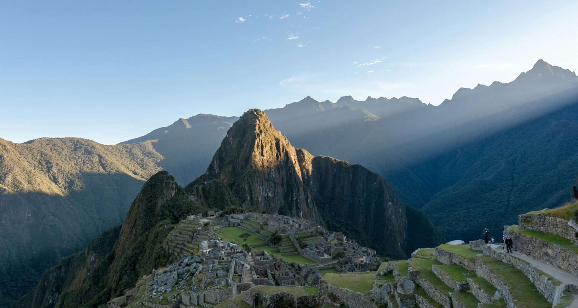 A scenic view of Machu Picchu, an ancient Incan citadel located high in the Andes Mountains of Peru, with green terraces, rugged peaks, and a clear blue sky in the background.