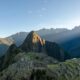 A scenic view of Machu Picchu, an ancient Incan citadel located high in the Andes Mountains of Peru, with green terraces, rugged peaks, and a clear blue sky in the background.