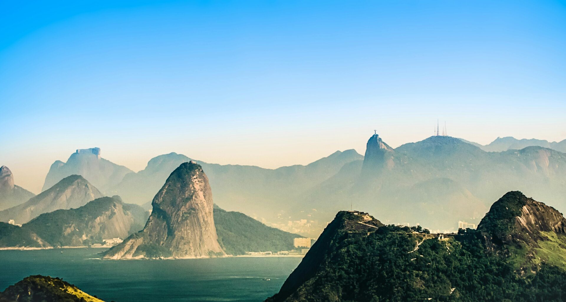 A breathtaking aerial view of Rio de Janeiro, showcasing its iconic mountains, lush greenery, and the serene coastline under a clear blue sky.